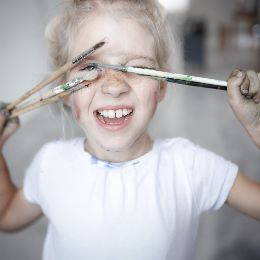 01_blondes-kleines-madchen-verspielter-stimmung-mit-farbe-auf-ihrem-sommersprossigen-gesicht-und-blauen-augen-die-ihr-gesicht-mit-pinseln-bedecken